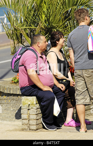 Un uomo in sovrappeso con la sua famiglia si siede su una parete e si prende una pausa dal camminare Foto Stock