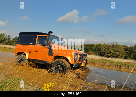 Colore arancio Land Rover Defender 90, dotato di un alto sollevato di aspirazione dell'aria, attraversando una foresta allagata via. Foto Stock