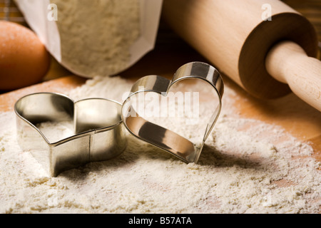 A forma di cuore ad cookie cutters in farina, mattarello, pack di farina, uova in background Foto Stock