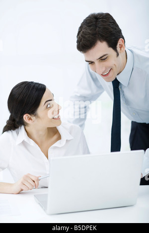 Professional donna seduta con il computer portatile, collega di sesso maschile guardando sopra la sua spalla, entrambi sorridente Foto Stock