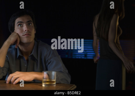 Uomo seduto vicino a bicchiere di whiskey, guardando in alto, woman dancing in background Foto Stock