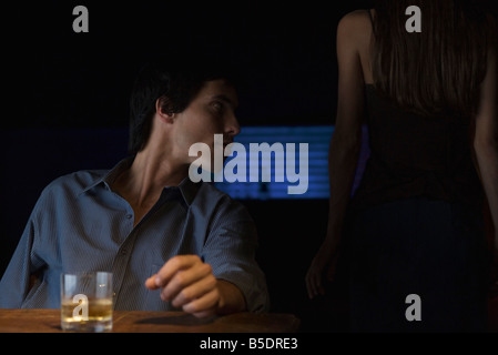 Bicchiere di whiskey su tavola, uomo seduto e guardando lontano, vista posteriore della donna in piedi nel buio Foto Stock