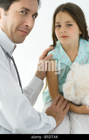 Pediatra consolante bambina, sorridente sulla spalla in telecamera Foto Stock