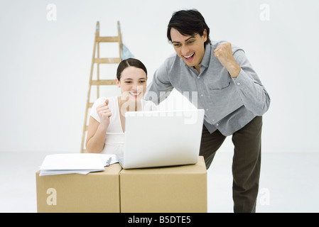 Coppia giovane guardando il computer portatile sulla parte superiore della scatola di cartone, sorridente, clenching a suon di pugni Foto Stock