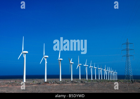 Le turbine eoliche che conduce al pilone di elettricità sulla costa soleggiata con cielo blu dietro Foto Stock