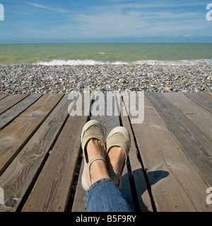 Vista di due gambe e piedi rilassante su una scheda camminare accanto a una spiaggia di ciottoli e oceano Foto Stock