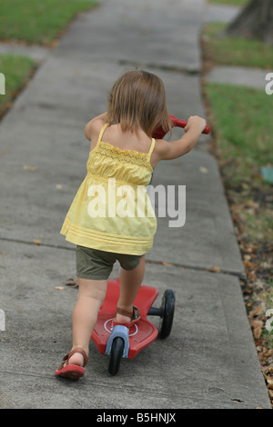 Un 2-anno-vecchia ragazza toddler cavalca il suo scooter giù suo quartiere marciapiede. Foto Stock