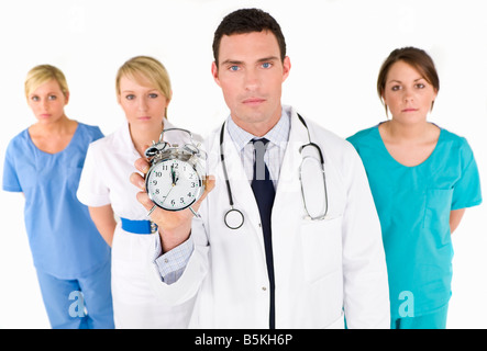 Un medico di sesso maschile sostenuto dal suo team medico tenendo fuori un allarme orologio che scandisce sempre più vicina alle ore 12. Focus sulla superficie dell'orologio. Foto Stock