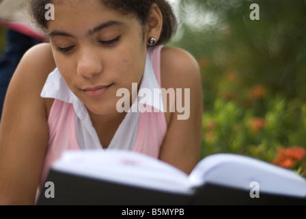 Un giovane adolescente Latina la lettura di un libro nero mentre recante su di un parco. Foto Stock