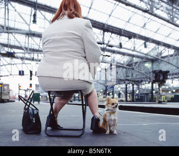 Il colore di una immagine a colori di un sovrappeso donna seduta su una piega fuori sgabello in una stazione ferroviaria con la sua chihuahua cane giocattolo Foto Stock