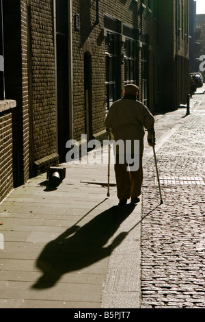 Silhouette e ombra di uomo anziano che indossa il tappo piatto a piedi lungo la strada di ciottoli con le stampelle Southwark Londra Foto Stock