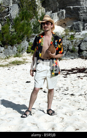 Giovane uomo che indossa Bermuda, una camicia hawaiana e un cappello di paglia in possesso di una noce di cocco, sulla spiaggia Stonehole, Bermuda Foto Stock