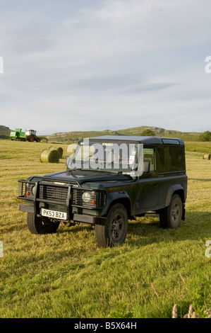 Un Land Rover Defender parcheggiato in un campo di fieno rotoli, mentre un trattore funziona in background. Foto Stock