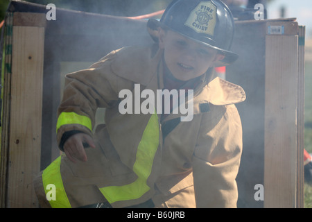 Un piccolo ragazzo che indossa una camicia firemens e fire chief hat a un incendio fiera sicurezza facendo un esercizio di formazione Foto Stock