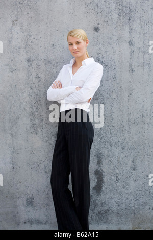 Giovane donna bionda che indossa abiti business in piedi con le braccia conserte di fronte a un muro grigio Foto Stock