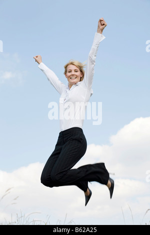 Giovane donna bionda che indossa abbigliamento business jumping davanti a un cielo blu, le braccia tese Foto Stock