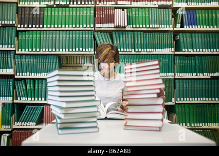 Giovani dai capelli scuri studente femmina seduto con molti libri a un tavolo di fronte a uno scaffale in una libreria Foto Stock