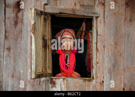 Giovane donna di Akha Pala tribù guardando fuori della finestra un vestito in un colorato testa-vestito di rosso e un necklace, Ban Saenkham T Foto Stock