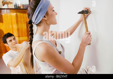 Giovane donna martellamento chiodo nel muro con un giovane uomo con una cornice immagine Foto Stock