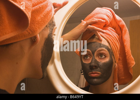 Giovane donna con la spugnetta esfoliante per il viso maschera guardando a se stessa e a specchio Foto Stock