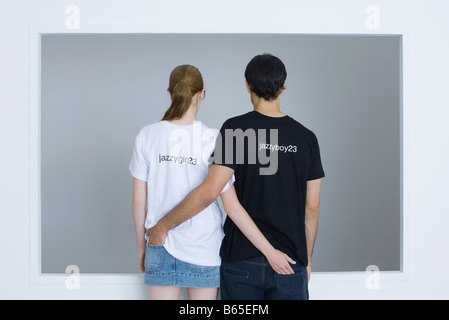 Coppia giovane indossando personalizzato tee-shirt, braccia intorno reciprocamente le cinture, vista posteriore Foto Stock