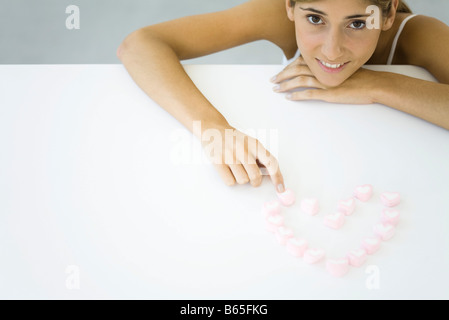 Donna con la caramella cuori disposti a forma di cuore, appoggiando il mento sulla mano, sorridente in telecamera Foto Stock