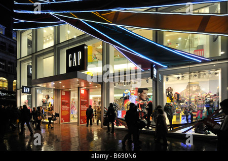 Decorazioni natalizie sera vista notturna degli acquirenti a piedi marciapiedi fuori dal negozio DI MODA GAP in un trafficato Oxford Street West End Londra Inghilterra UK Foto Stock