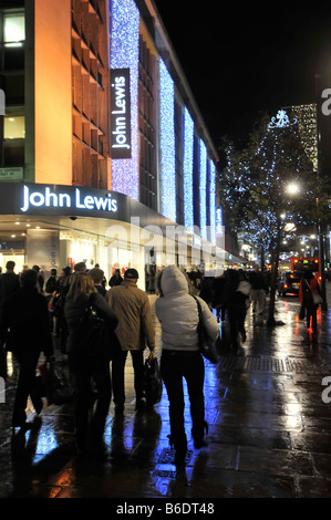 Gli acquirenti che camminano su marciapiedi bagnati John Lewis grande magazzino Oxford Street a Londra West End con luci di strada di Natale e decorazioni Inghilterra Regno Unito Foto Stock