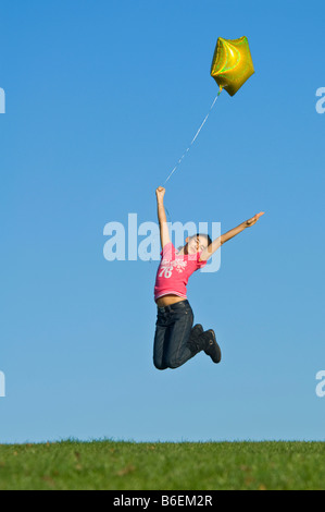 Una giovane ragazza (11) saltare in aria con un giallo elio palloncino riempito contro un cielo blu. Foto Stock