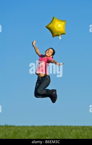 Una giovane ragazza (11) saltare in aria con un giallo elio palloncino riempito contro un cielo blu. Foto Stock