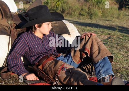 Un giovane cowboy in appoggio contro il suo letto a rullo e slitta sulla gamma dopo una dura giornata di lavoro Foto Stock