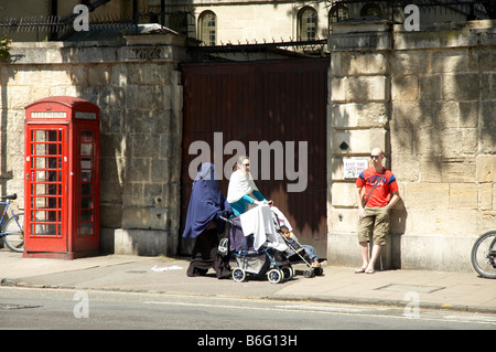 Due donne, una con abiti occidentali e una con un velo da niqab, camminano con un passeggino davanti a una cabina telefonica rossa a High Street, Oxford, Regno Unito. Foto Stock