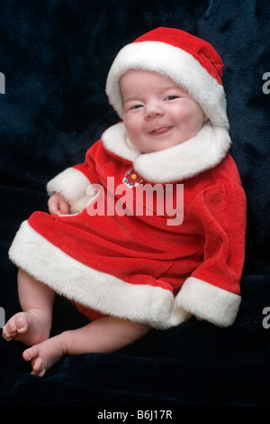 Tre mesi ragazza sorridente, vestito in un abito di Santa e cappello di Natale per contro uno sfondo scuro Foto Stock