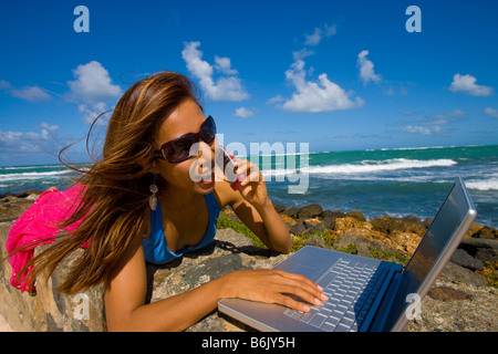 Giovane donna asiatica usando un computer portatile presso la spiaggia mentre sta parlando su un telefono cellulare, San Juan, Puerto Rico Foto Stock