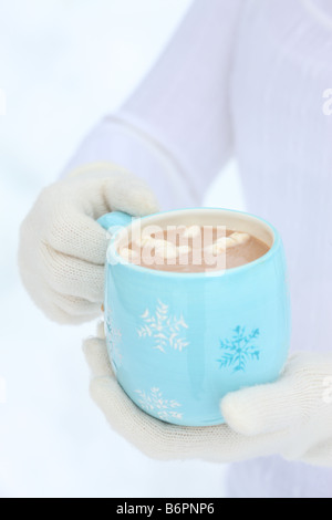 Le mani con guanti invernali tenendo premuto il simbolo del fiocco di neve blu mug con cioccolata calda e marshmallows Foto Stock