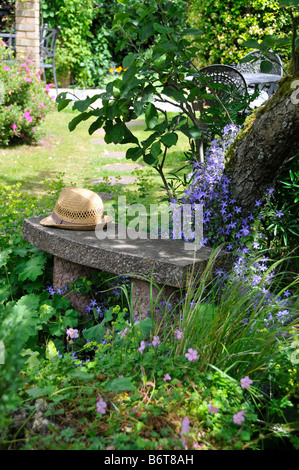 Tradizionale giardino inglese scena con cappello di paglia sul banco sotto albero circondato da fiori Foto Stock