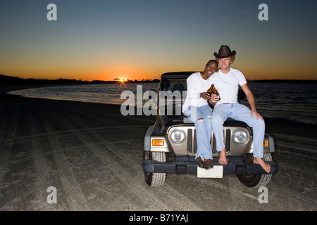 Giovani interracial giovane seduto su una jeep a beach, tostatura bottiglia di birra, tramonto Foto Stock