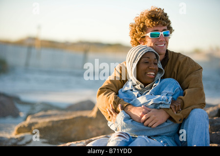 Giovani interracial giovane seduto sulla spiaggia con braccio intorno a Foto Stock