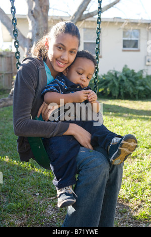 African American pre-teen ragazza seduta sul mantenimento in rotazione il fratello del bambino nel cortile posteriore Foto Stock