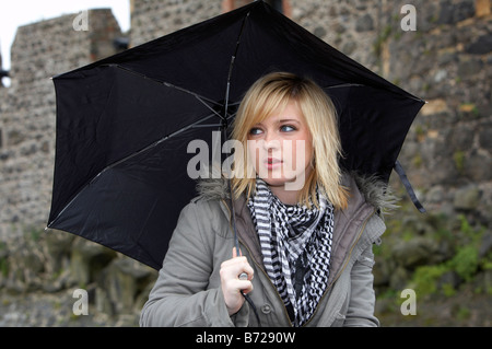 Blonde 18 enne ragazza con ombrello in giacca e sciarpa pesante guardando lontano dalla fotocamera nella parte anteriore Carrickfergus Castle Foto Stock