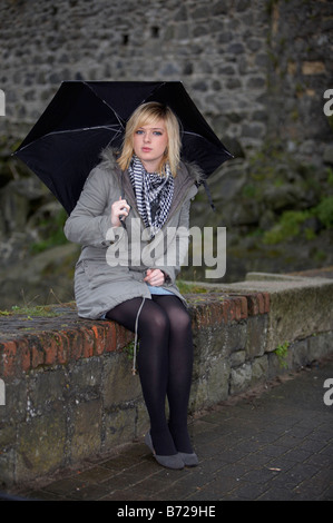 Blonde 18 enne ragazza con ombrello nero in giacca e sciarpa pesante contatto visivo seduto sul muro basso Foto Stock