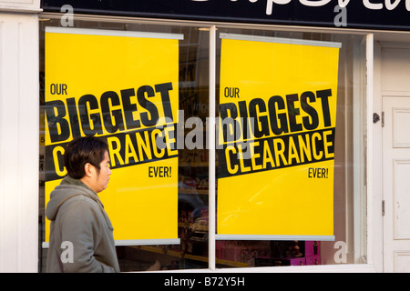 2 giallo vendita segni in una finestra del negozio sostenendo il gioco più grande con una donna SHOPPER PASSATO A PIEDI Foto Stock