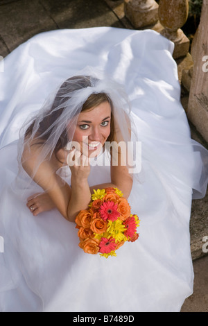 Giovane e bella sposa in abito da sposa al di fuori seduta con mazzo di fiori Foto Stock