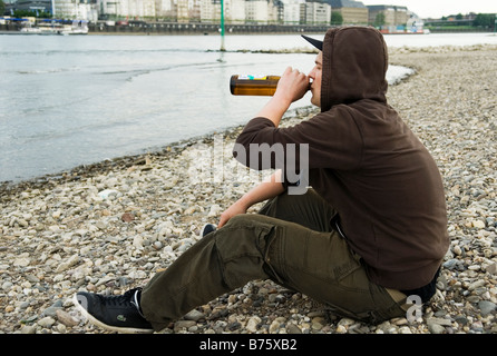 Lonely ragazzo adolescente si siede sul lungofiume e bevande birra fuori di una bottiglia e fuma una sigaretta Foto Stock