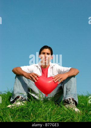 16-anno-vecchio ragazzo seduto su un prato, tenendo un cuore palloncino a forma di Foto Stock