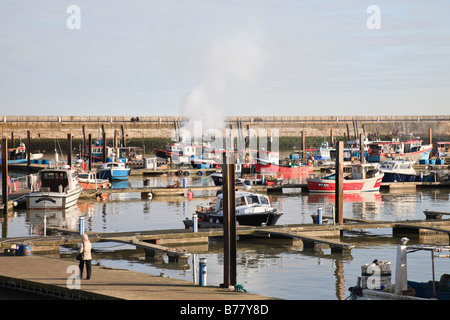 Una donna cammina lungo un pontone verso barche da pesca ormeggiate a Ramsgate Royal Harbour, uno ha del fumo proveniente dal suo camino Foto Stock