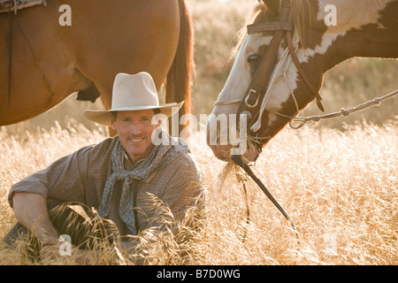 Un cowboy seduto in erba lunga con due cavalli Foto Stock