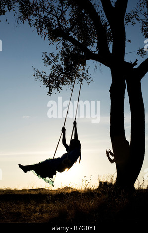 Silhouette di ragazze indiane basculante in una oscillazione in casa nella rurale campagna indiana al tramonto Foto Stock