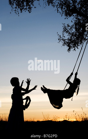 Silhouette di ragazze indiane basculante in una oscillazione in casa nella rurale campagna indiana al tramonto. Andhra Pradesh, India Foto Stock