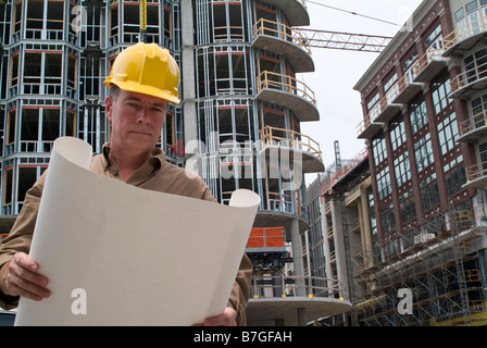Un uomo in un hardhat guardando un grande pezzo di carta con la nuova costruzione in background Foto Stock